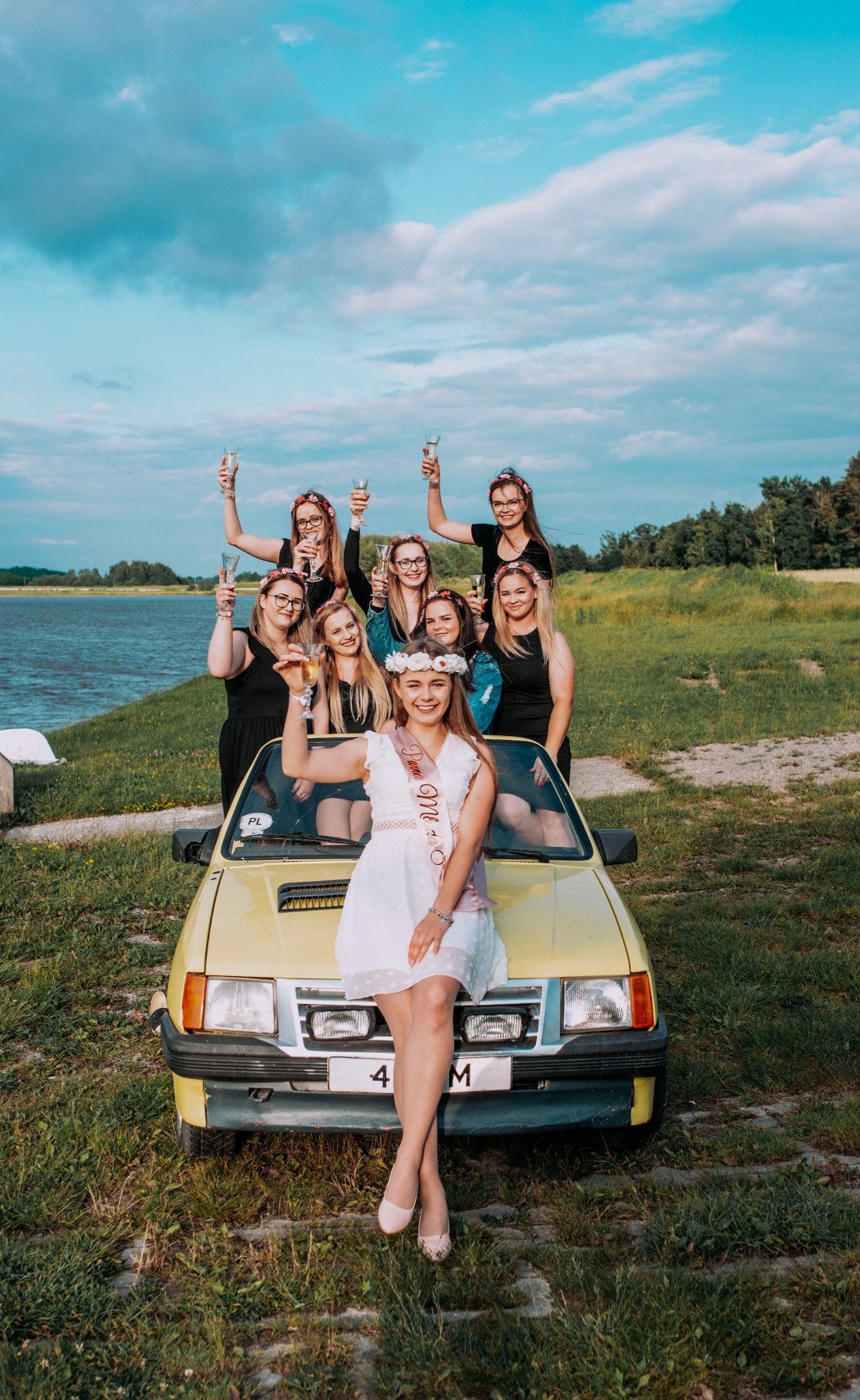 Grupa kobiet świętuje wieczór panieński, stojąc i siedząc na żółtym samochodzie na tle jeziora i pochmurnego nieba, trzymając kieliszki z szampanem.