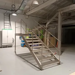 Metalowe schody przemysłowe z platformą, wykonane ze stali nierdzewnej, prowadzące do wyższego poziomu hali produkcyjnej, z widocznymi instalacjami pod sufitem i elementami wyposażenia hali w tle.