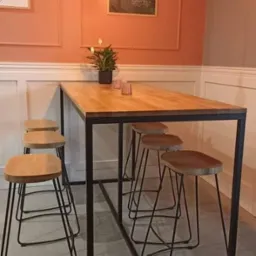 Wysoki stół z blatem z jasnego drewna i czarną metalową ramą, otoczony drewnianymi stołkami barowymi na metalowych nogach, w tle ściana w odcieniach różu i beżu z dekoracjami.