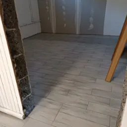 Ułożona podłoga z płytek imitujących drewno, widok przez fragment odnowionego wejścia z surową ścianą i białą boazerią.