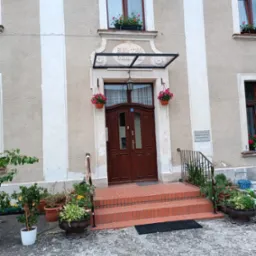 Wejście do budynku z ceglanymi schodami, balustradą z ciemnego metalu i kwiatami w donicach, nad drzwiami zadaszenie z napisem 'BUD. 1790 ODN. 1978'.
