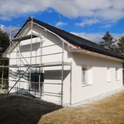 Świeżo otynkowana elewacja domu jednorodzinnego z zamaskowanymi oknami i rusztowaniem przy ścianie szczytowej, przygotowana do malowania.