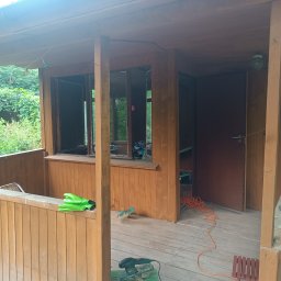 Taras Prytula.UA🇺🇦 - Drewniana altana w trakcie renowacji: widoczne narzędzia, szlifierka, przedłużacz, rękawice robocze, oraz rosnąca roślina na podłodze, sugerująca konieczność pielęgnacji otoczenia.