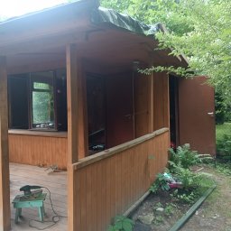 Taras Prytula.UA🇺🇦 - Drewniana altana ogrodowa z otwartymi drzwiami, widoczne narzędzia do obróbki drewna na zewnątrz, zielone rękawice i roślinność wokół