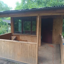 Taras Prytula.UA🇺🇦 - Drewniana altana z oknami i werandą, widok z zewnątrz, częściowo pomalowana, z rozłożonym kablem elektrycznym na podłodze.