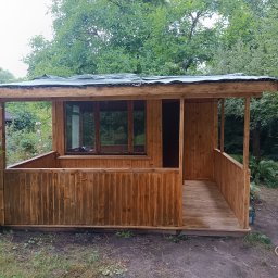 Taras Prytula.UA🇺🇦 - Drewniana altana ogrodowa z oknami i zadaszeniem z blachy falistej, otoczona zielenią drzew i krzewów, widok z przodu.