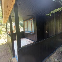 Taras Prytula.UA🇺🇦 - Zadaszony taras drewniany po świeżym malowaniu na czarno, okna zabezpieczone folią malarską, widoczne ślady farby na podłożu.