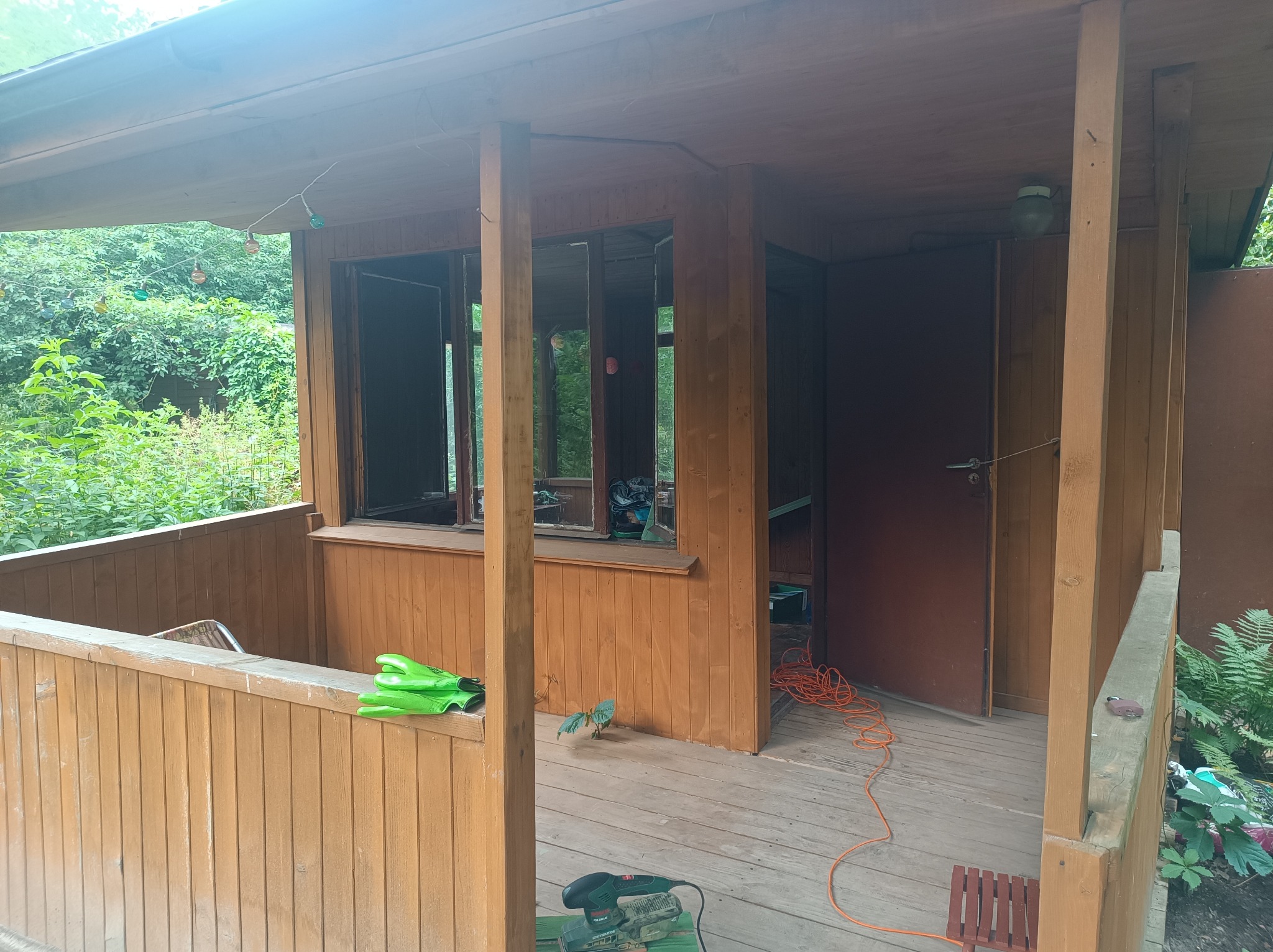 Drewniana altana w trakcie renowacji: widoczne narzędzia, szlifierka, przedłużacz, rękawice robocze, oraz rosnąca roślina na podłodze, sugerująca konieczność pielęgnacji otoczenia.