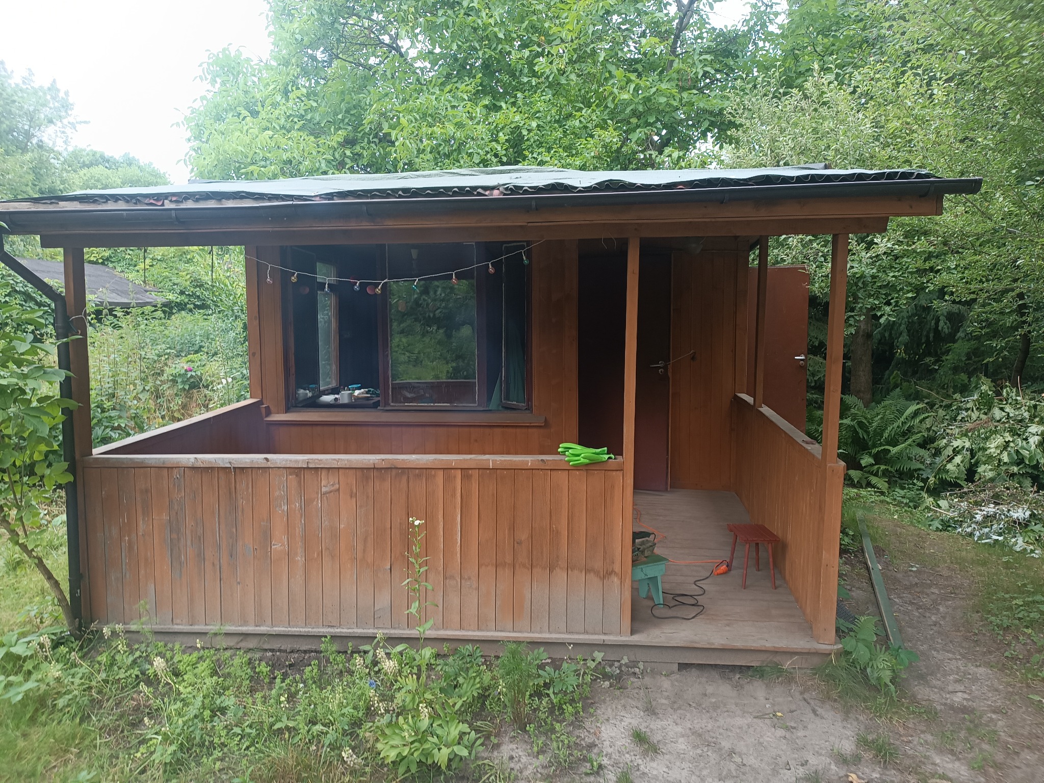 Drewniana altana z zadaszeniem, otoczona zielenią, z widocznymi narzędziami ogrodniczymi i stołkiem. W oknach zawieszone ozdobne lampki.