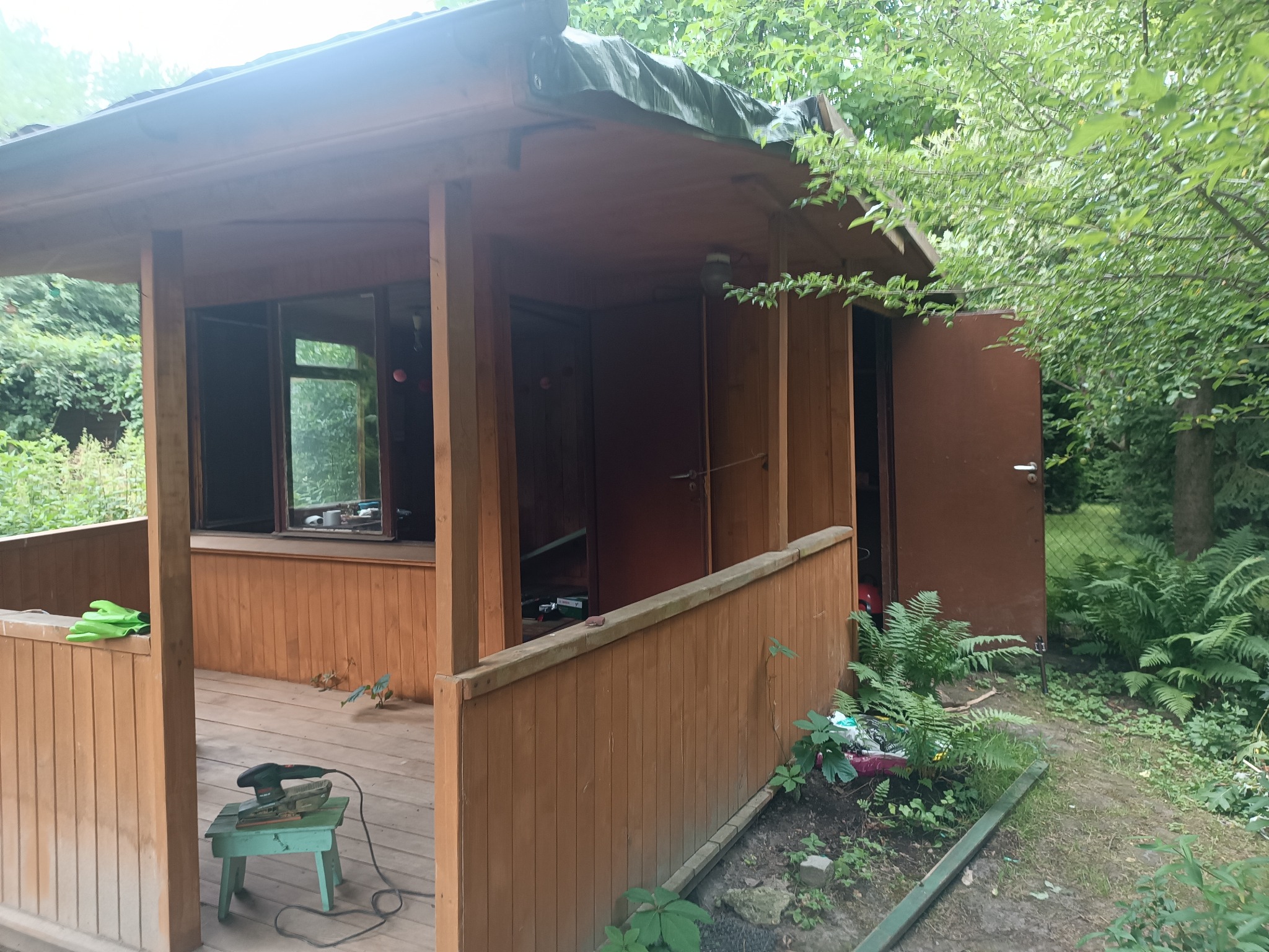 Drewniana altana ogrodowa z otwartymi drzwiami, widoczne narzędzia do obróbki drewna na zewnątrz, zielone rękawice i roślinność wokół