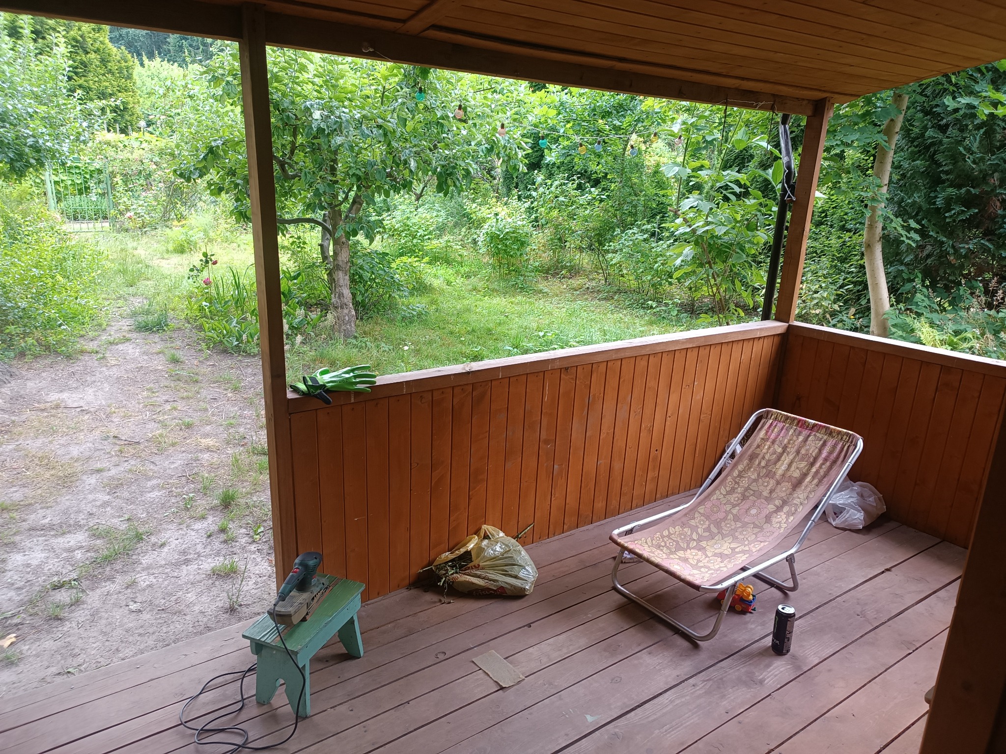 Zadaszony taras z drewnianą podłogą i widokiem na ogród, leżak plażowy, szlifierka na małym taborecie, worek z odpadami ogrodowymi i rękawice robocze.