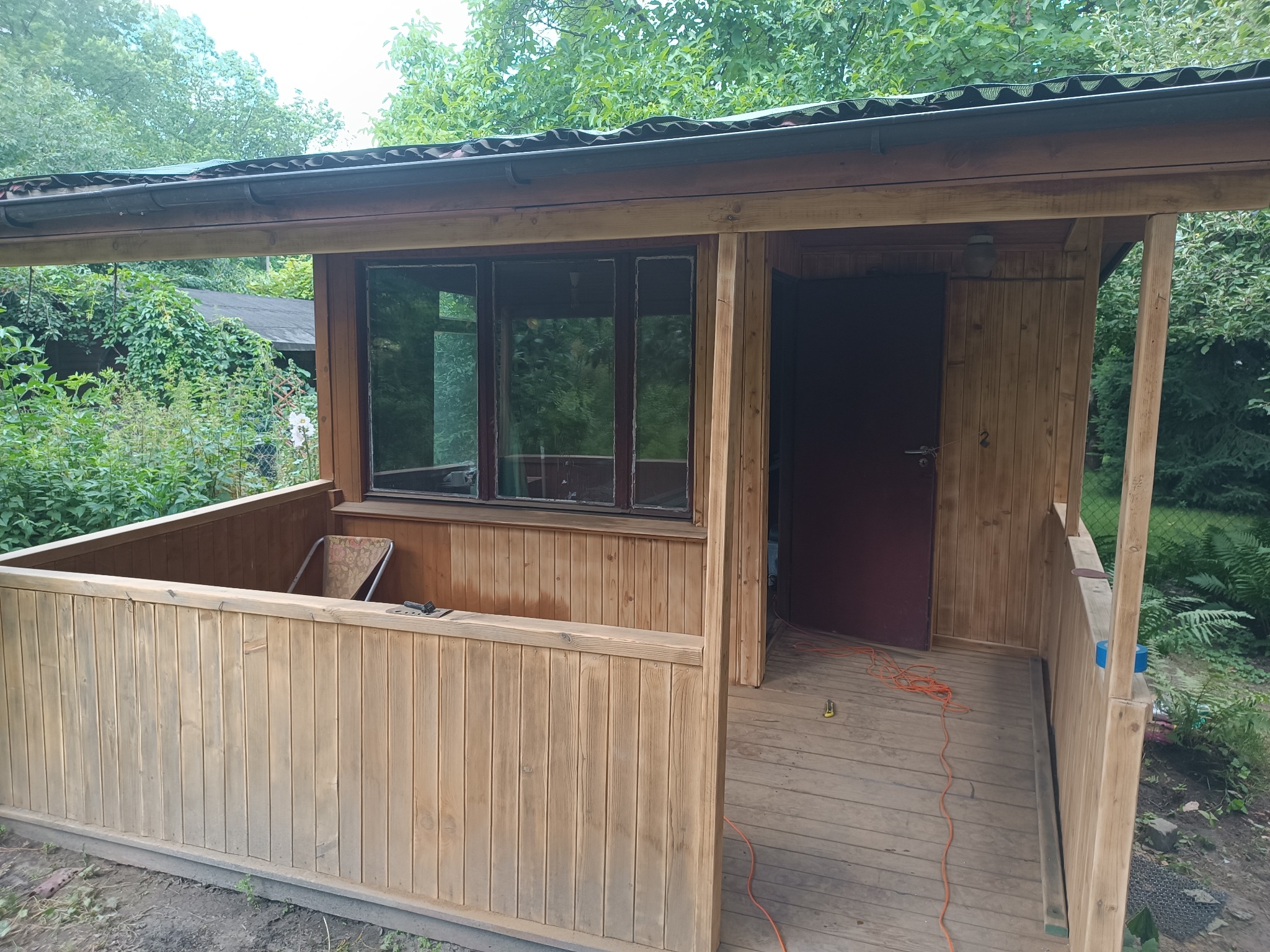 Drewniana altana z oknami i werandą, widok z zewnątrz, częściowo pomalowana, z rozłożonym kablem elektrycznym na podłodze.