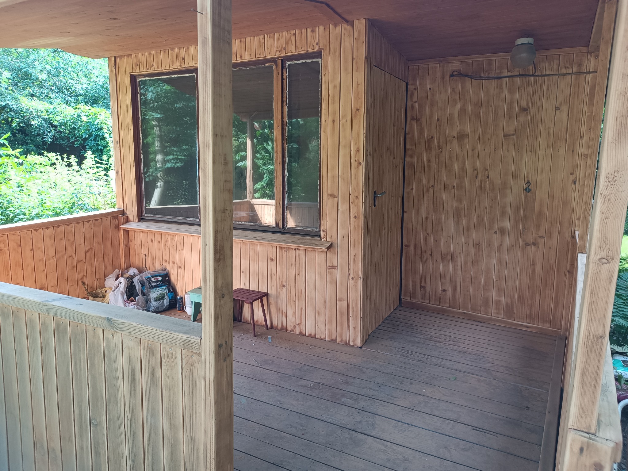 Zadaszony, drewniany taras w Warszawie z widokiem na zieleń, wyposażony w małe okna, drewniane balustrady i prosty, drewniany stołek. W rogu tarasu widoczne worki z ziemią i narzędzia ogrodnicze.