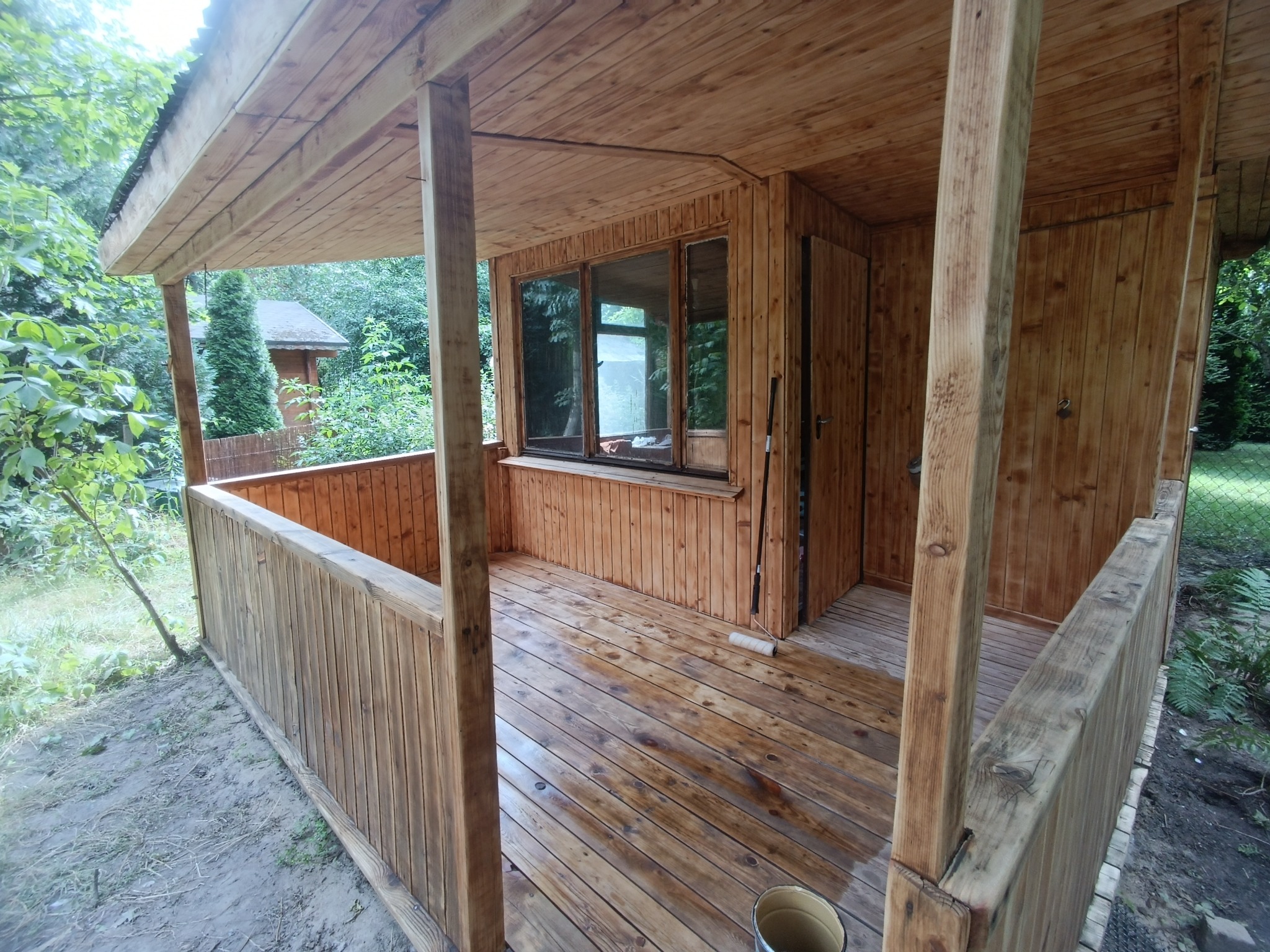 Drewniana altana z widokiem na ogród, świeżo pomalowana podłoga z widocznymi smugami mokrej farby, wałek malarski oparty o ścianę, wiadro w rogu, zieleń za oknami.