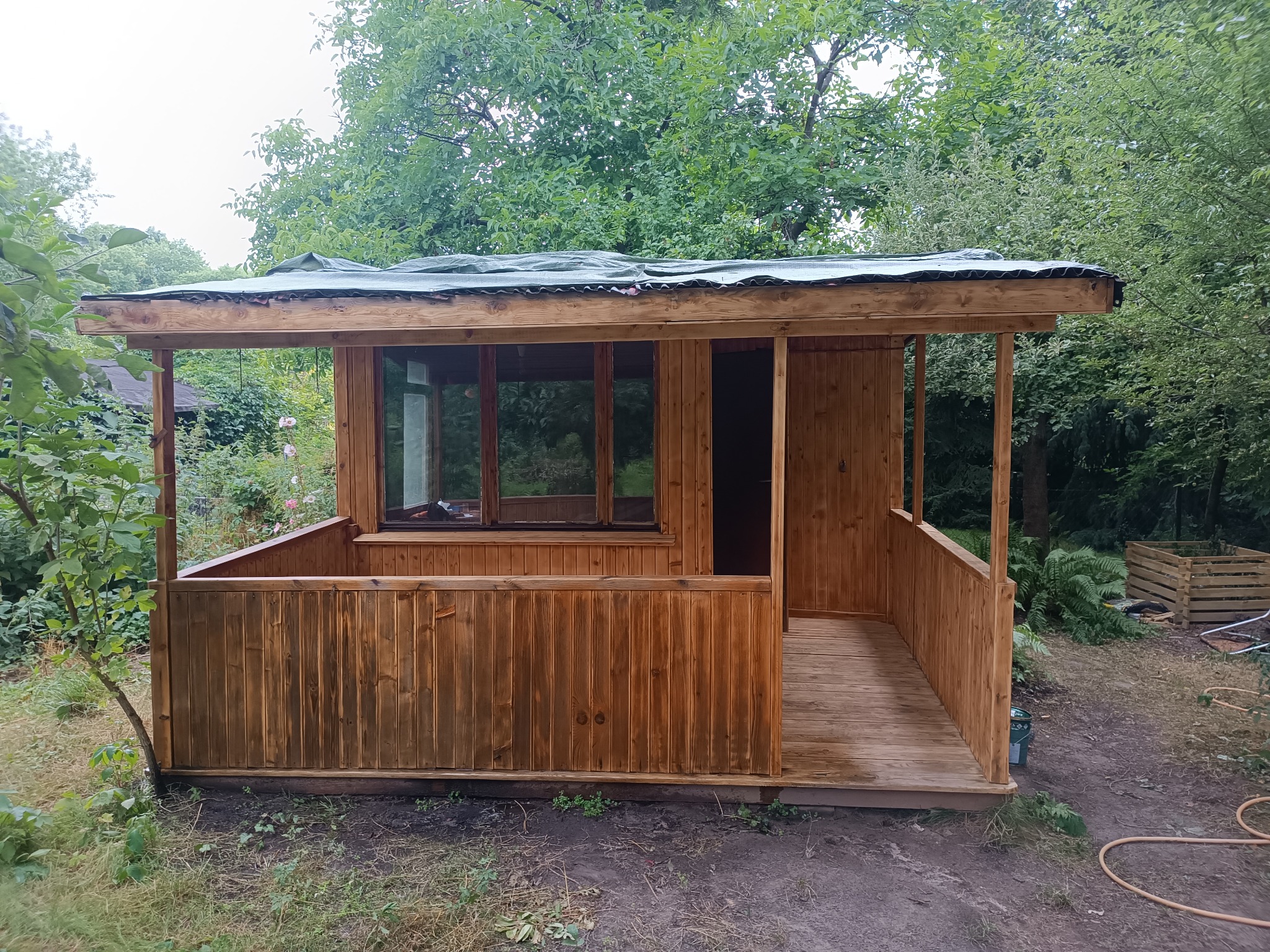 Drewniana altana ogrodowa z oknami i zadaszeniem z blachy falistej, otoczona zielenią drzew i krzewów, widok z przodu.