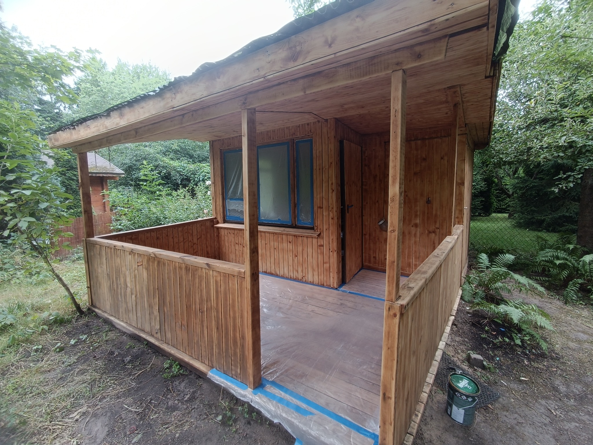 Drewniana altana ogrodowa po impregnacji, z folią ochronną na podłodze i zaklejoną taśmą malarską wokół okien, otoczona zielenią ogrodu.