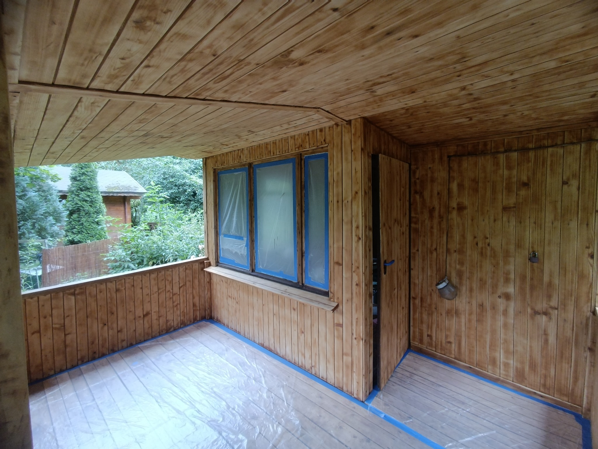 Drewniany taras w trakcie malowania bejcą, okna zabezpieczone folią, widoczne ślady bejcy na podłodze i ścianach.