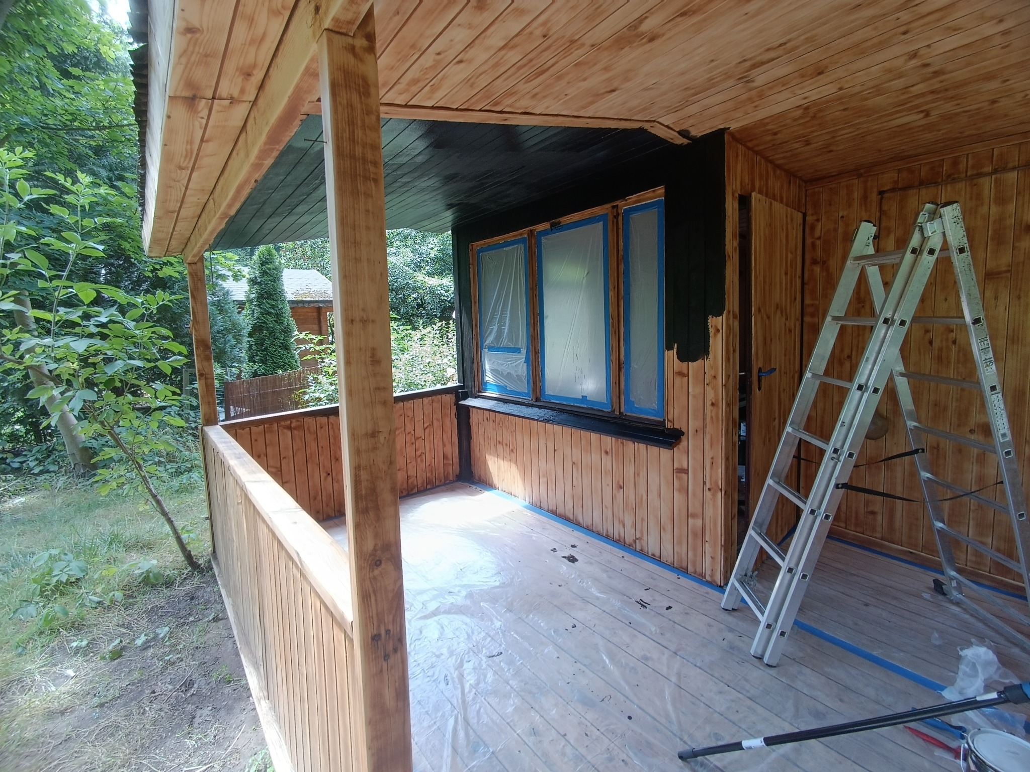 Malowanie drewnianej altany: widoczne zabezpieczone okna folią, świeżo pomalowany na czarno fragment ściany, drabina oparta o ścianę, podłoga zabezpieczona folią malarską, widoczne pędzle i wiadro...