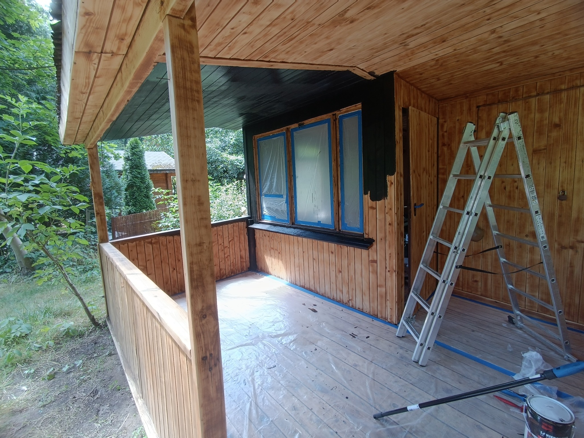 Malowanie drewnianej altany na działce w Warszawie: widoczna drabina, farba, zaklejone okna i pomalowana na czarno górna część ściany.