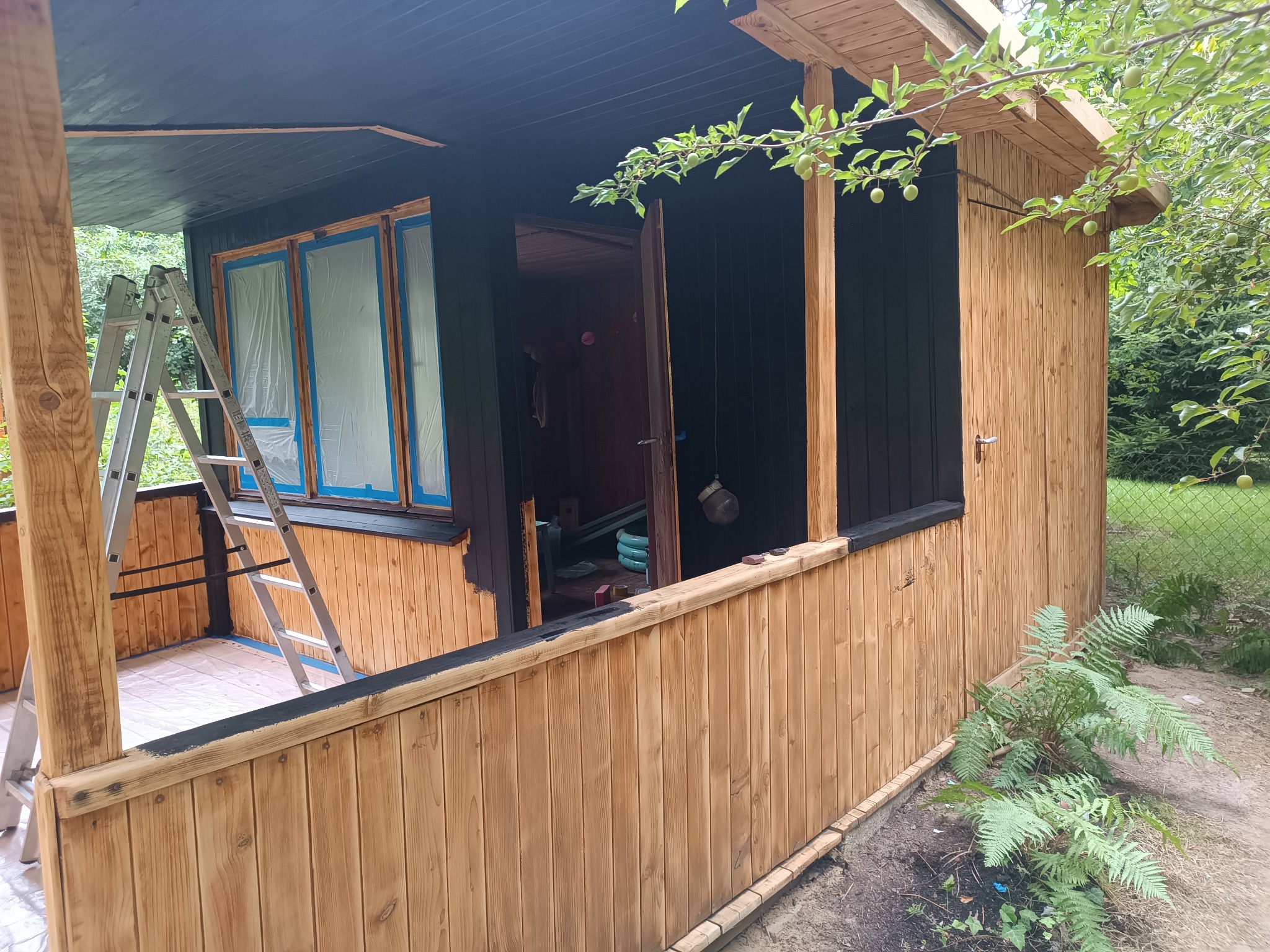Drewniana altana w trakcie malowania na czarno, okna zabezpieczone folią, drabina oparta o ścianę, widoczne elementy konstrukcyjne i fragment zieleni w tle.