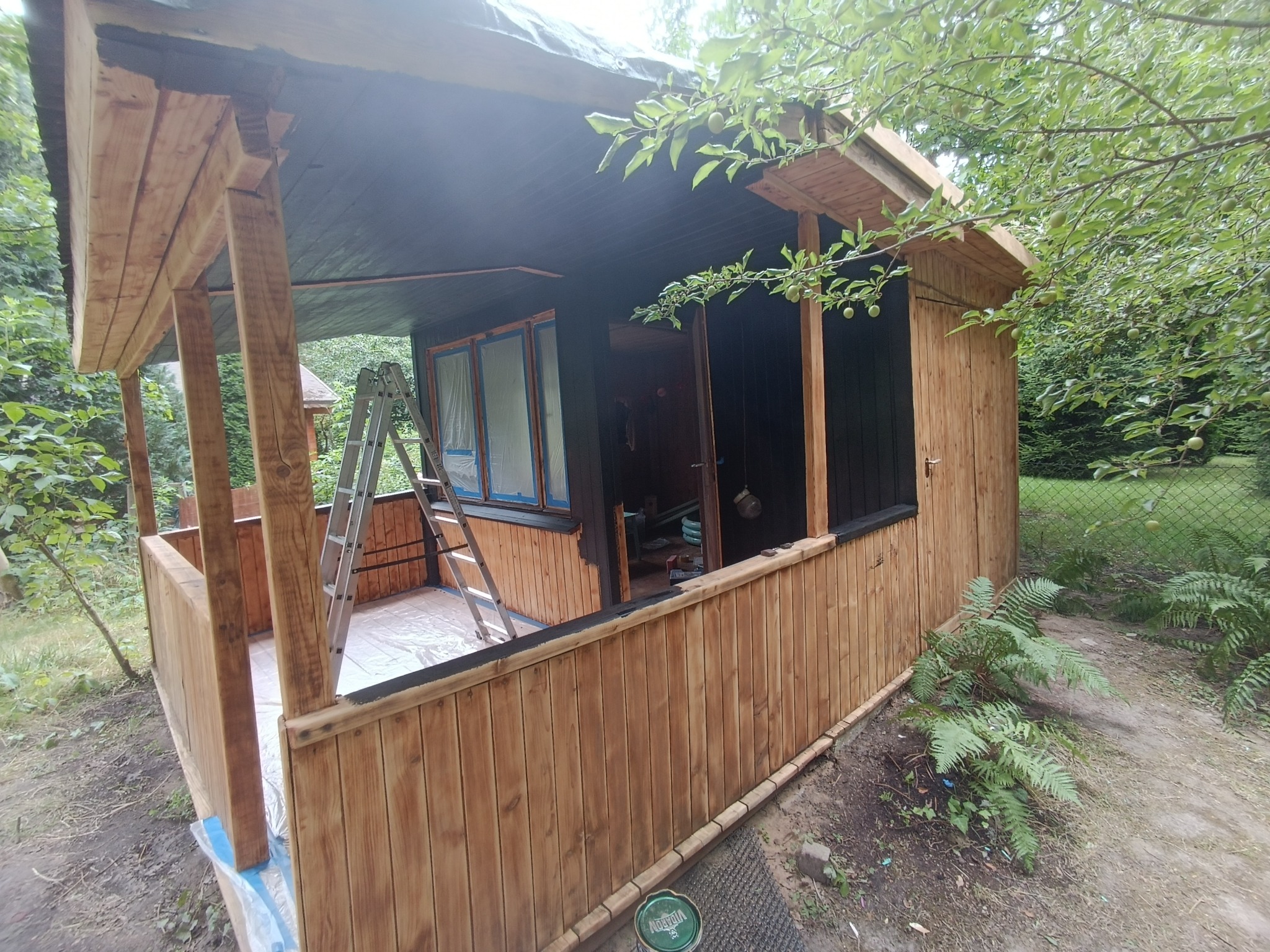 Malowanie drewnianej altany ogrodowej na czarno, widoczna drabina i zabezpieczona folią podłoga, otoczona zielenią.