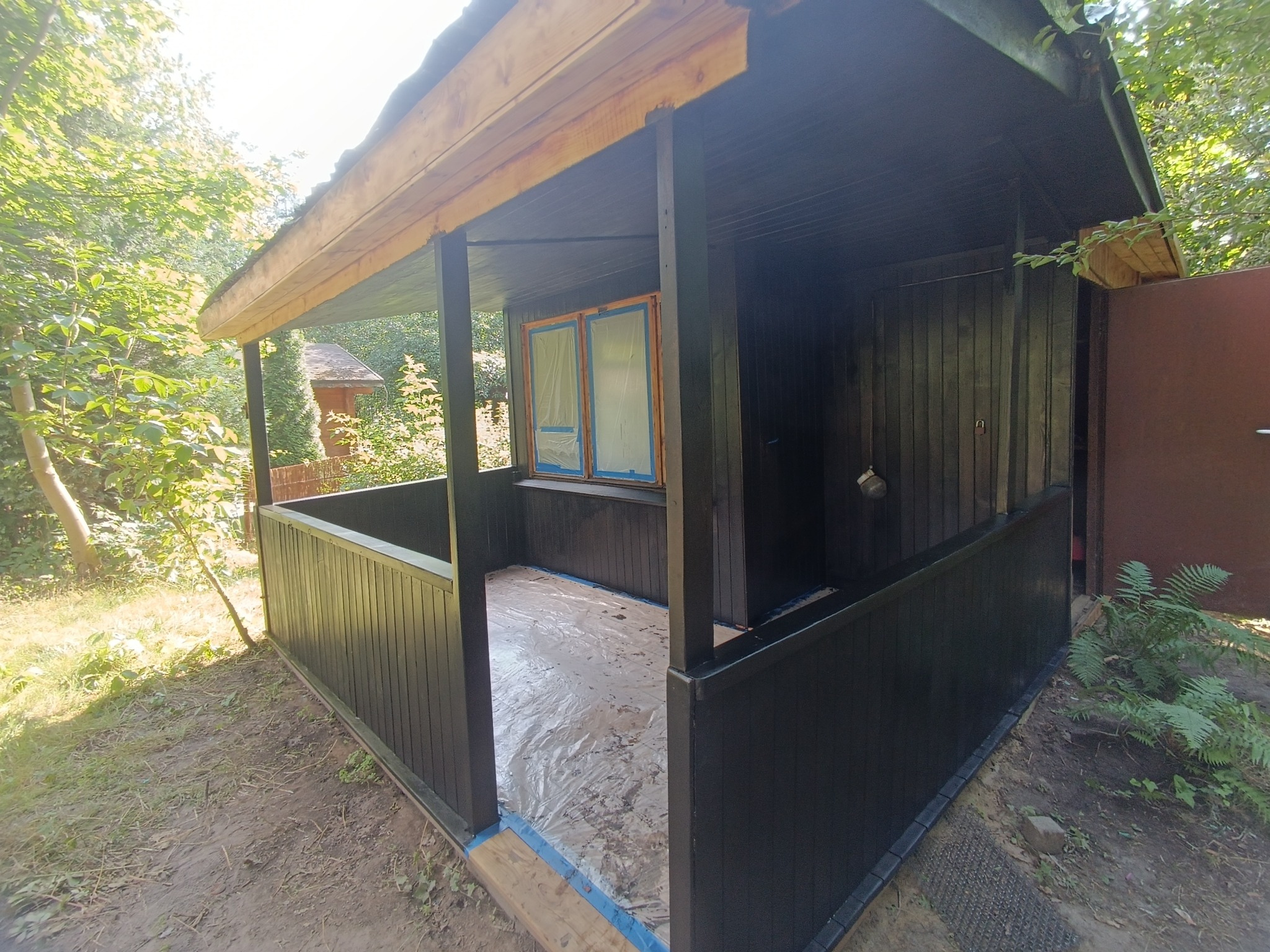 Świeżo pomalowana na czarno drewniana altana ogrodowa, z zabezpieczonymi folią oknami i podłogą, w otoczeniu zieleni.