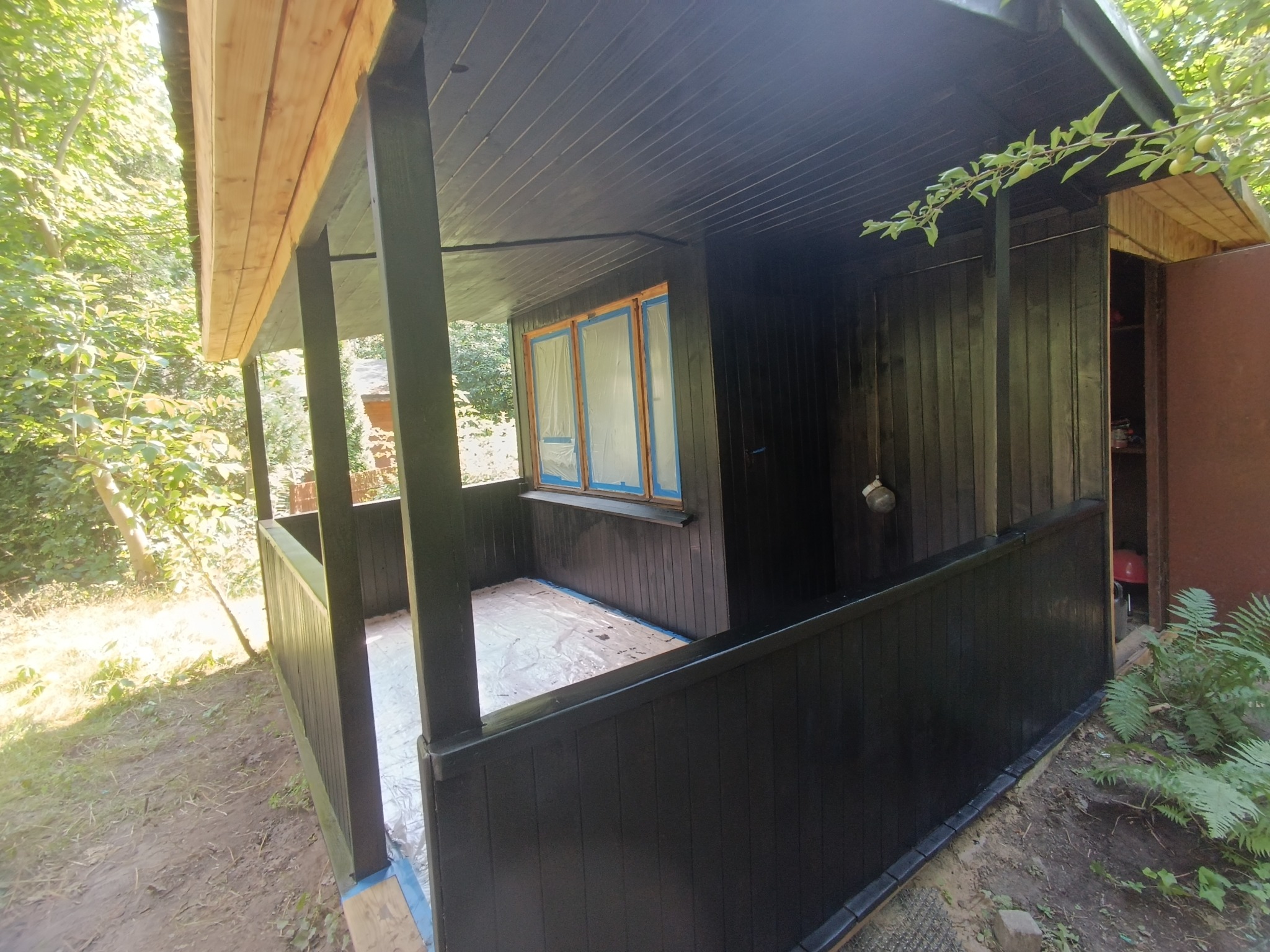 Zadaszony taras drewniany po świeżym malowaniu na czarno, okna zabezpieczone folią malarską, widoczne ślady farby na podłożu.
