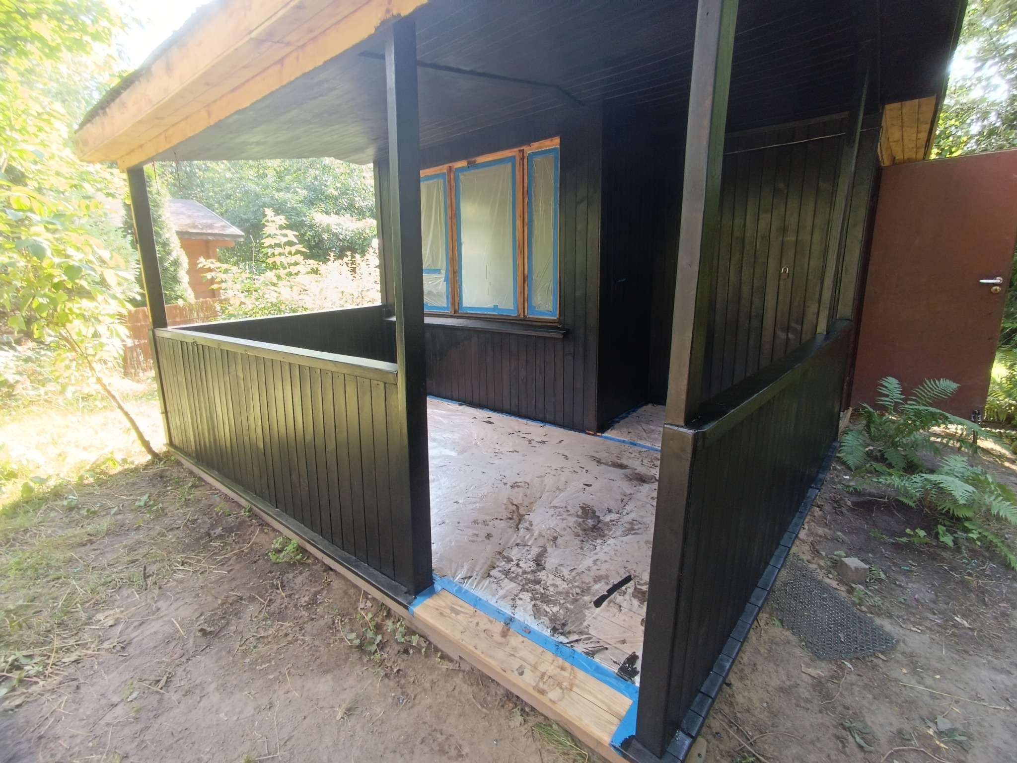 Malowanie altany ogrodowej na ciemny kolor, okna zabezpieczone folią malarską, podłoga przykryta folią ochronną, widoczne ślady farby.