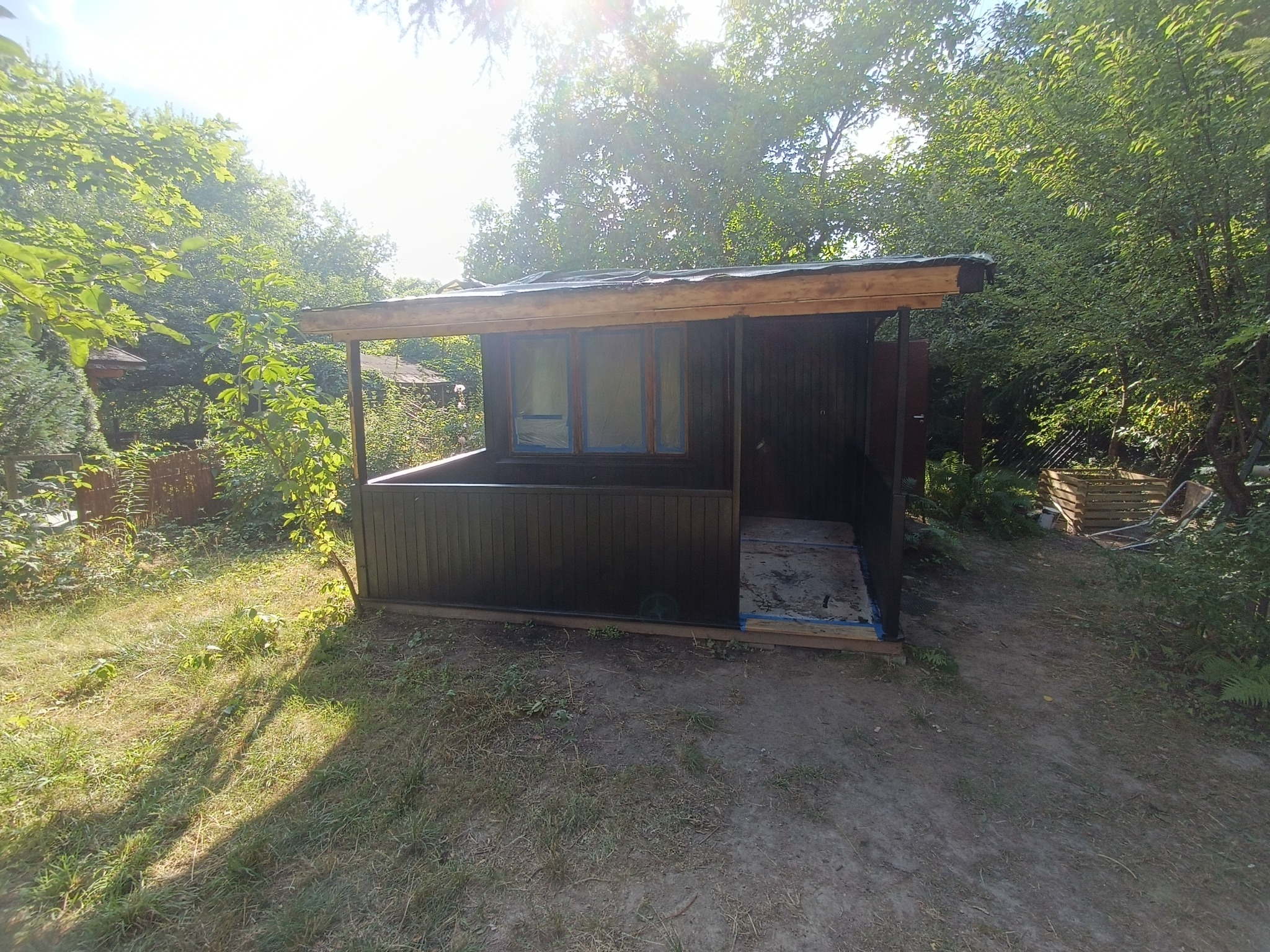 Nowo pomalowana, ciemnobrązowa altana ogrodowa z drewnianą konstrukcją, widoczna w słoneczny dzień, z taśmą malarską zabezpieczającą szyby, otoczona zielenią i drzewami.