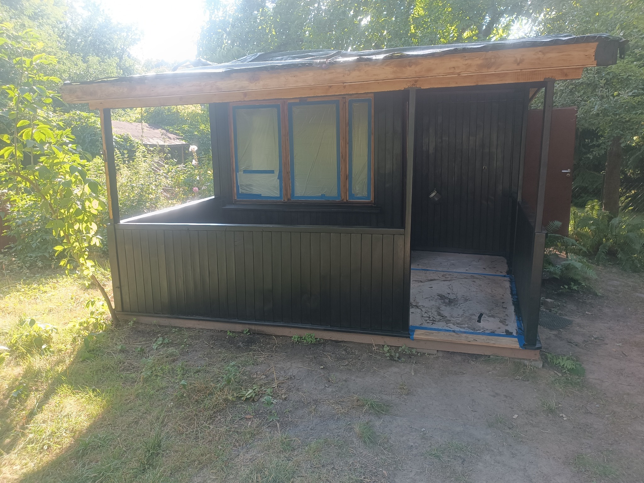Malowanie altany ogrodowej na kolor ciemny grafit, okna zabezpieczone folią i taśmą malarską, widok z zewnątrz na tle zieleni.