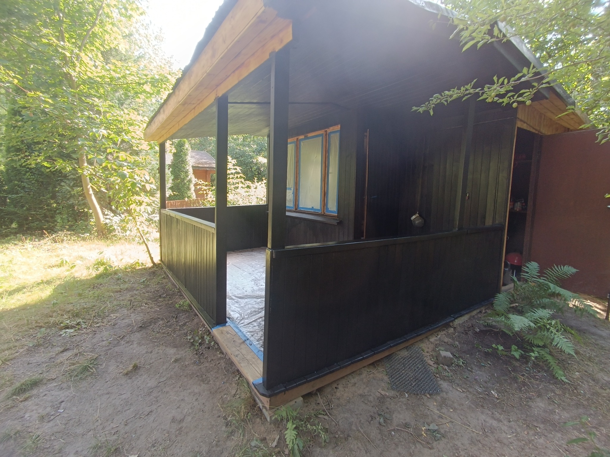 Ciemnobrązowa drewniana altana z widocznymi śladami świeżego malowania, zabezpieczona folią na podłodze, otoczona zielenią i drzewami.