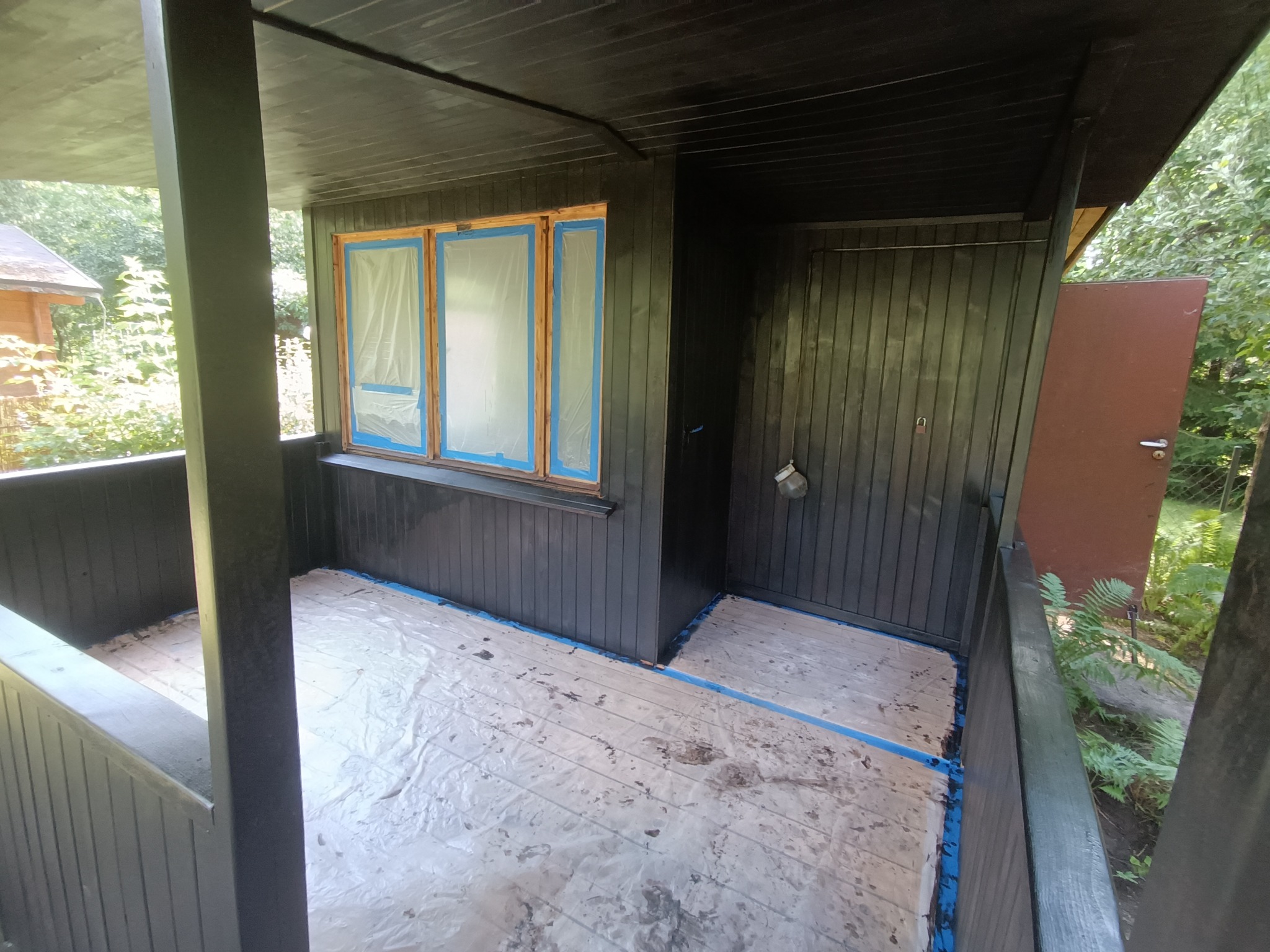 Zadaszony taras w trakcie malowania na ciemny kolor, okna zabezpieczone folią i taśmą malarską, podłoga przykryta folią ochronną przed zabrudzeniem farbą.