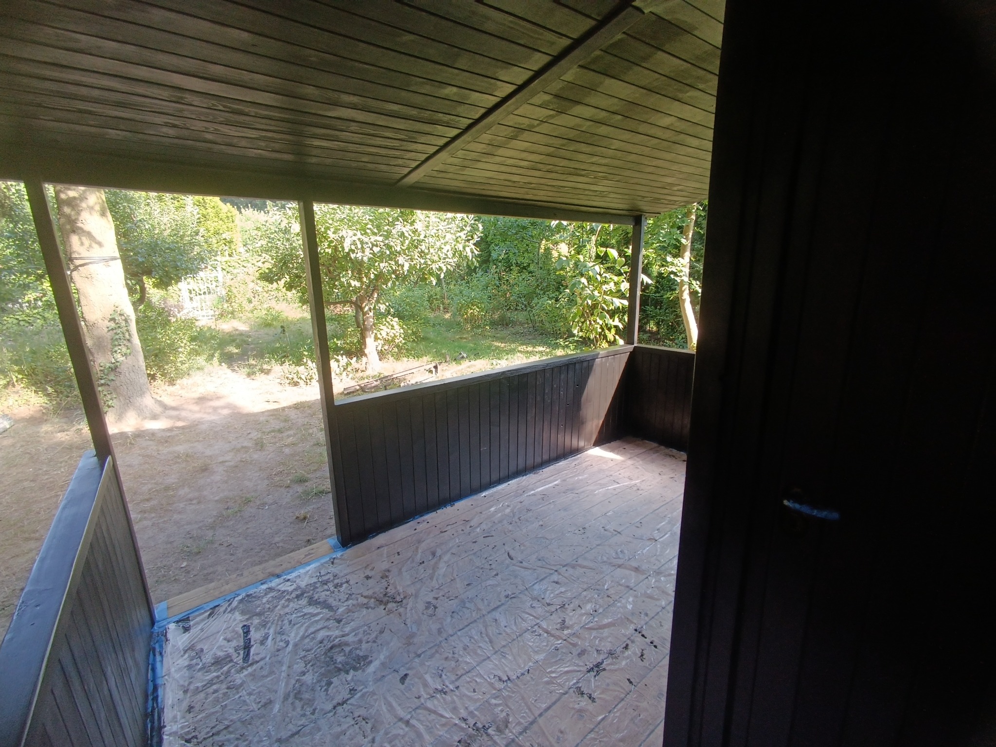 Malowanie drewnianej altany na ciemny kolor, podłoga zabezpieczona folią malarską, widok na ogród.