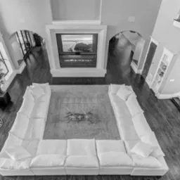 Przestronny salon z wysokim sufitem, widok z góry na białą sofę ustawioną w kształt litery U, nowoczesny kominek z telewizorem i sztucznym ogniem, ciemne drewniane podłogi i marmurowe wykończenia...