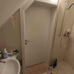 Doorway Krystian Stępień - Białe drzwi wewnętrzne z klamką w łazience z kabiną prysznicową, widok na umywalkę i kosmetyki.