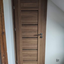 Doorway Krystian Stępień - Zamknięte, nowoczesne drzwi wewnętrzne z drewnianym fornirem i poziomymi, czarnymi akcentami, zamontowane w pokoju z białymi ścianami i drewnianą podłogą, widoczny fragment drewnianej belki w tle.