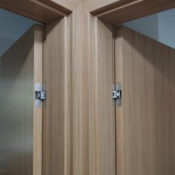 Doorway Krystian Stępień - Dwie drewniane ościeżnice drzwiowe z ukrytymi zawiasami, połączone pod kątem w rogu pomieszczenia.