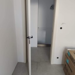 Doorway Krystian Stępień - Otwarta biała rama drzwiowa z czarną klamką, widok na białe wnętrze z kartonami w rogu, ślady po instalacji elektrycznej na ścianie.