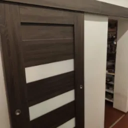 Ciemnobrązowe drzwi przesuwne z poziomymi, białymi wstawkami, zamontowane w ścianie, z widoczną zawartością szafy w tle.