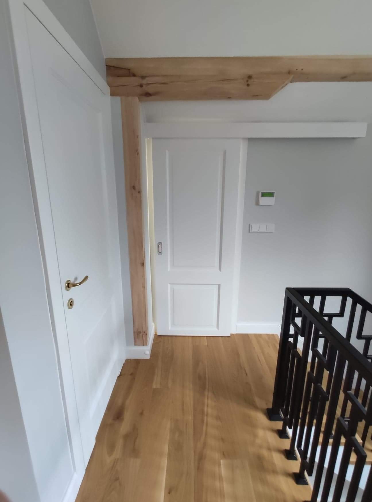 Białe, panelowe drzwi przesuwne z drewnianą ościeżnicą i widoczną prowadnicą, obok klasyczne drzwi z mosiężną klamką, parkiet na podłodze i czarna balustrada schodów.