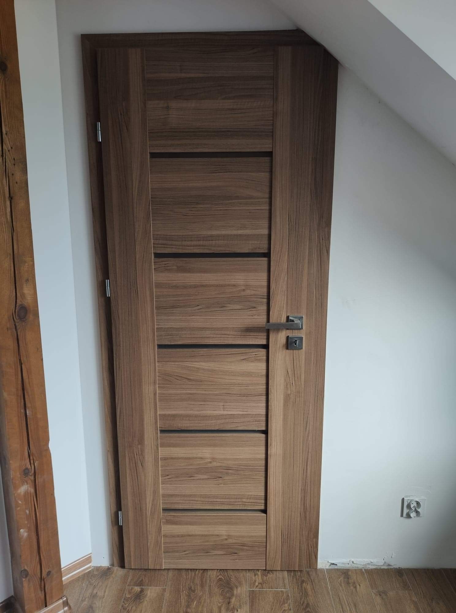 Zamknięte, nowoczesne drzwi wewnętrzne z drewnianym fornirem i poziomymi, czarnymi akcentami, zamontowane w pokoju z białymi ścianami i drewnianą podłogą, widoczny fragment drewnianej belki w tle.