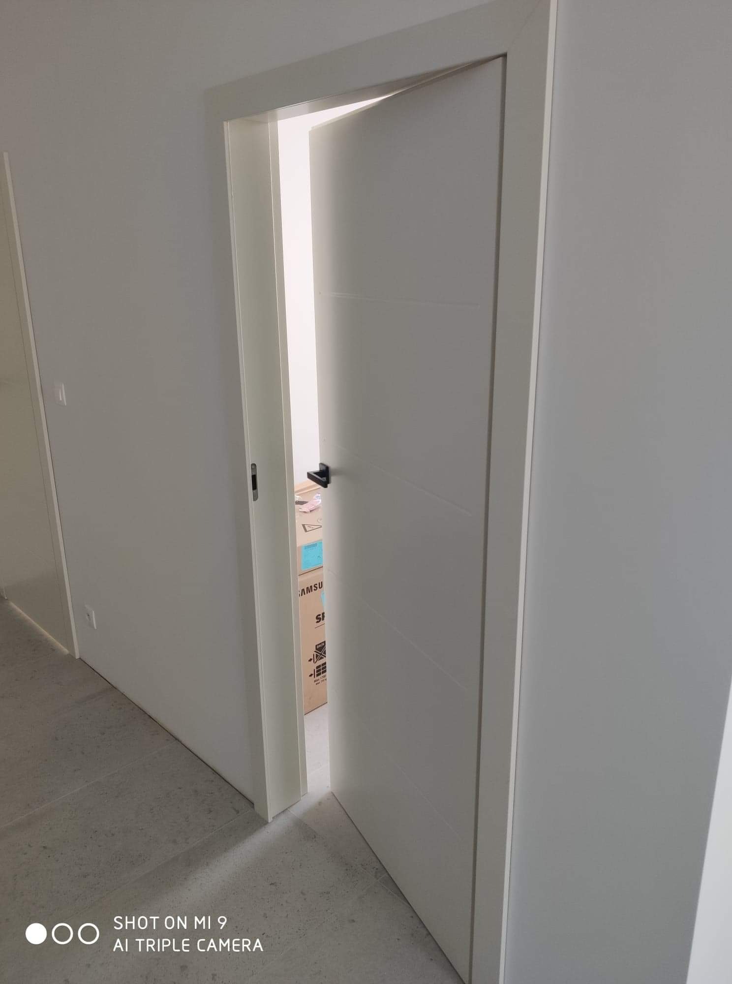 Otwarta, minimalistyczna biała brama z czarną klamką, widok na kartony w pomieszczeniu za nią, szara podłoga.