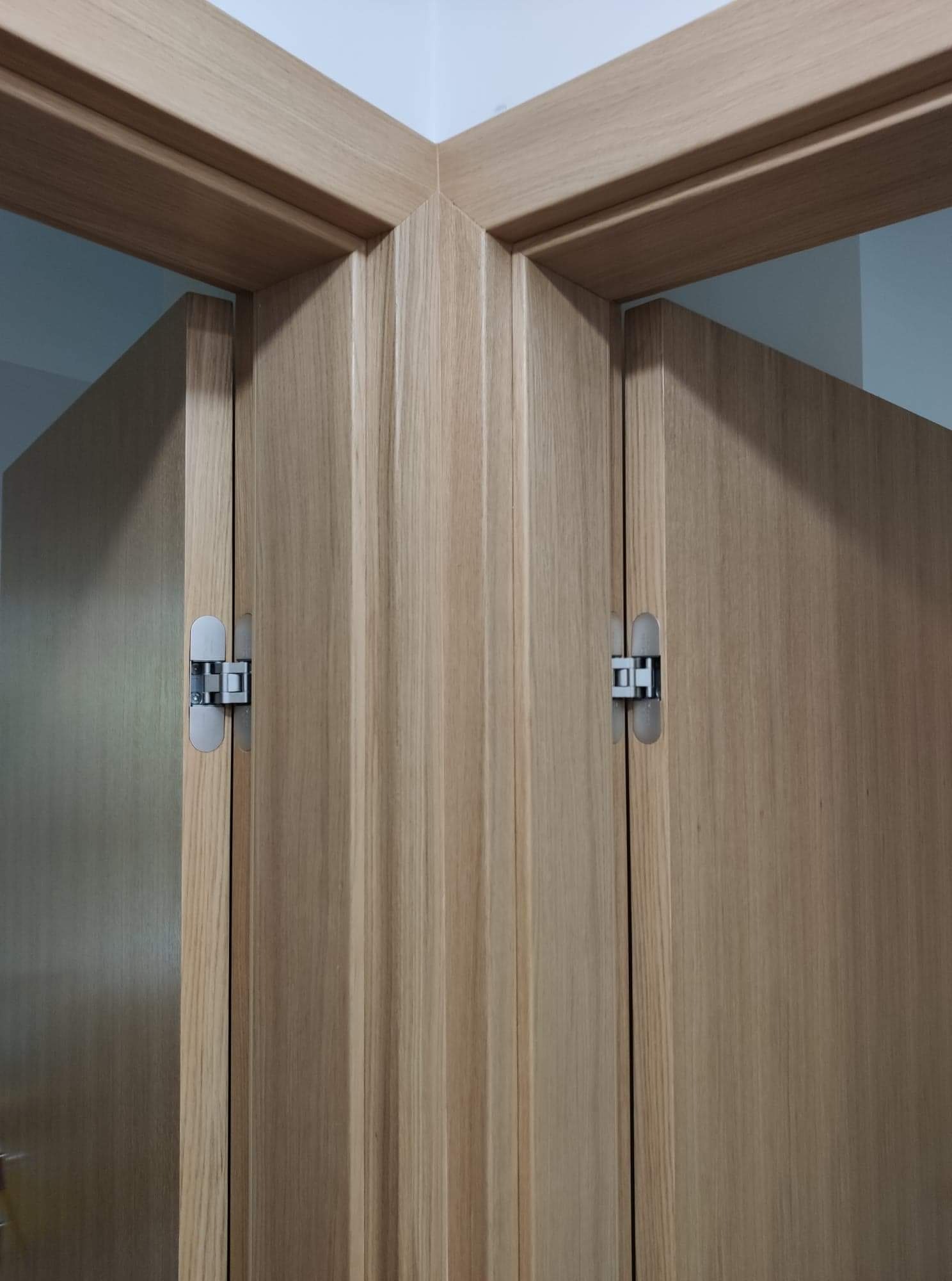 Dwie drewniane ościeżnice drzwiowe z ukrytymi zawiasami, połączone pod kątem w rogu pomieszczenia.