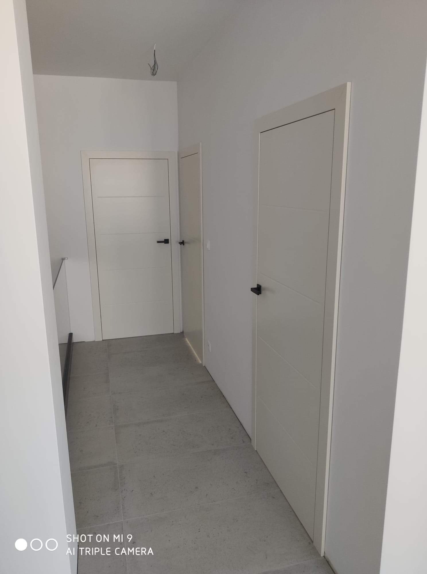 Dwa białe skrzydła drzwiowe z poziomymi frezowaniami i czarnymi klamkami w minimalistycznym korytarzu z szarą podłogą z płytek.