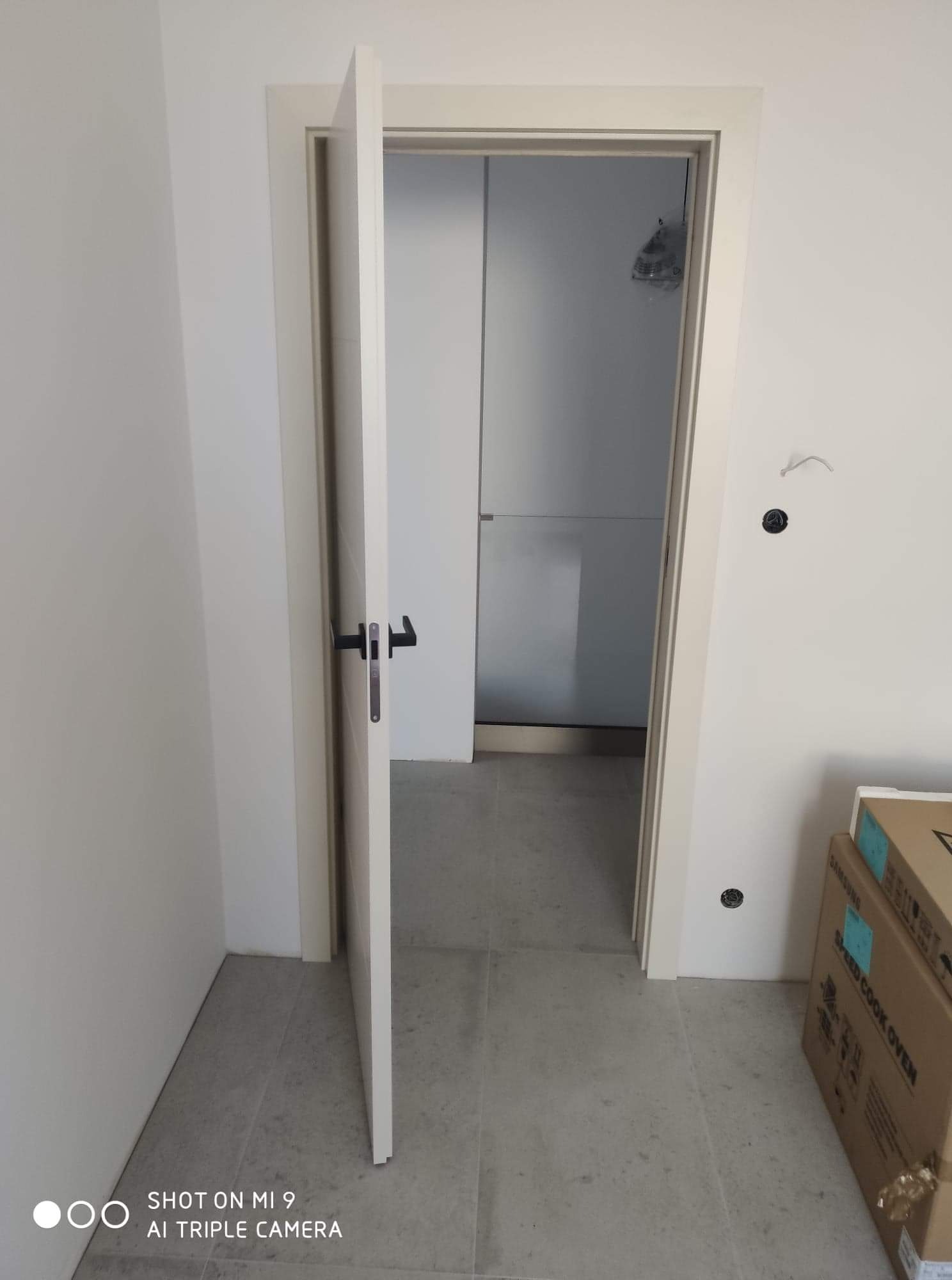 Otwarta biała rama drzwiowa z czarną klamką, widok na białe wnętrze z kartonami w rogu, ślady po instalacji elektrycznej na ścianie.