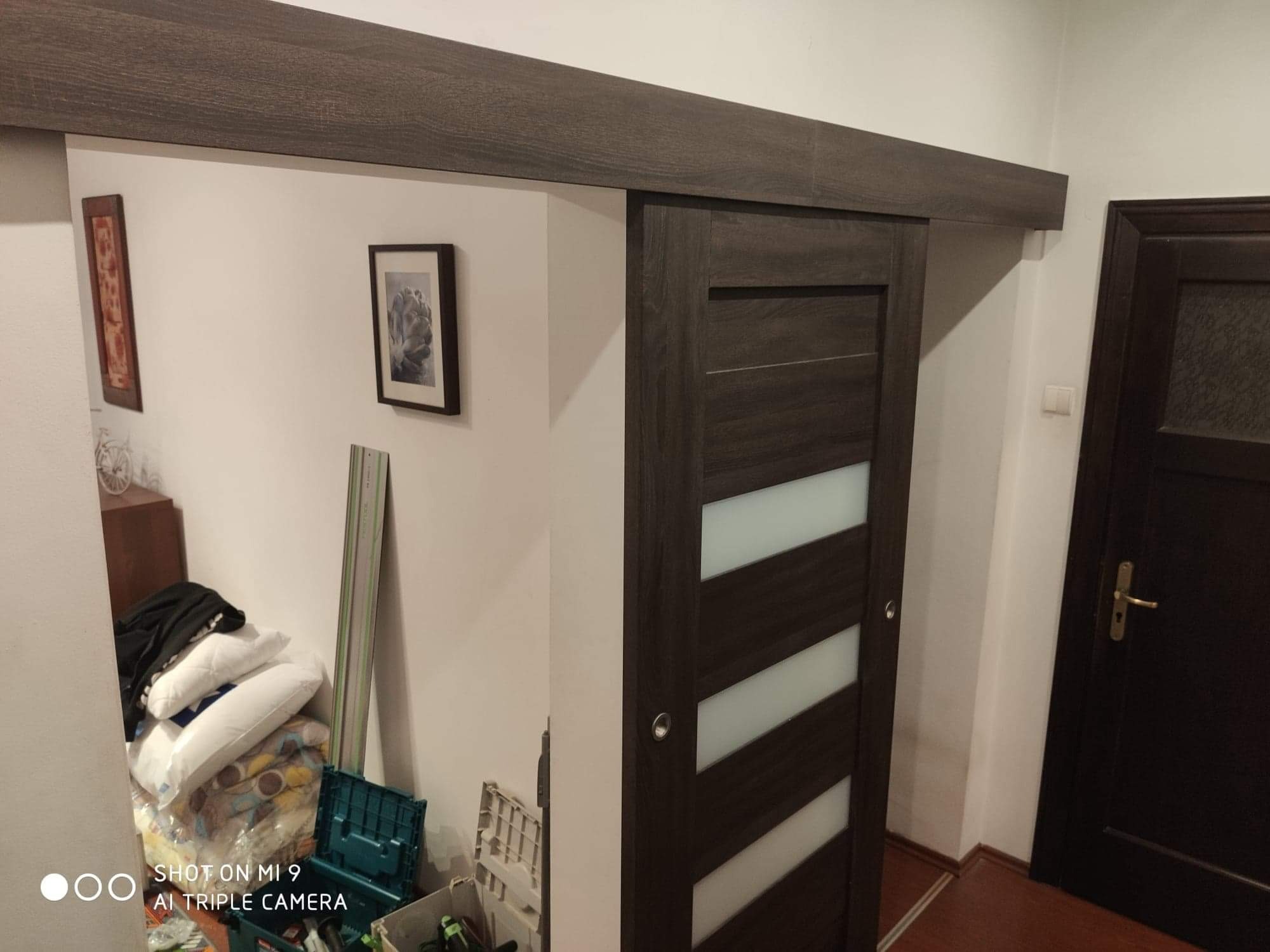 Zamontowane drzwi przesuwne z ciemnego drewna z poziomymi, matowymi paskami, widoczne w korytarzu z elementami wystroju wnętrz i drugimi drzwiami w tle.