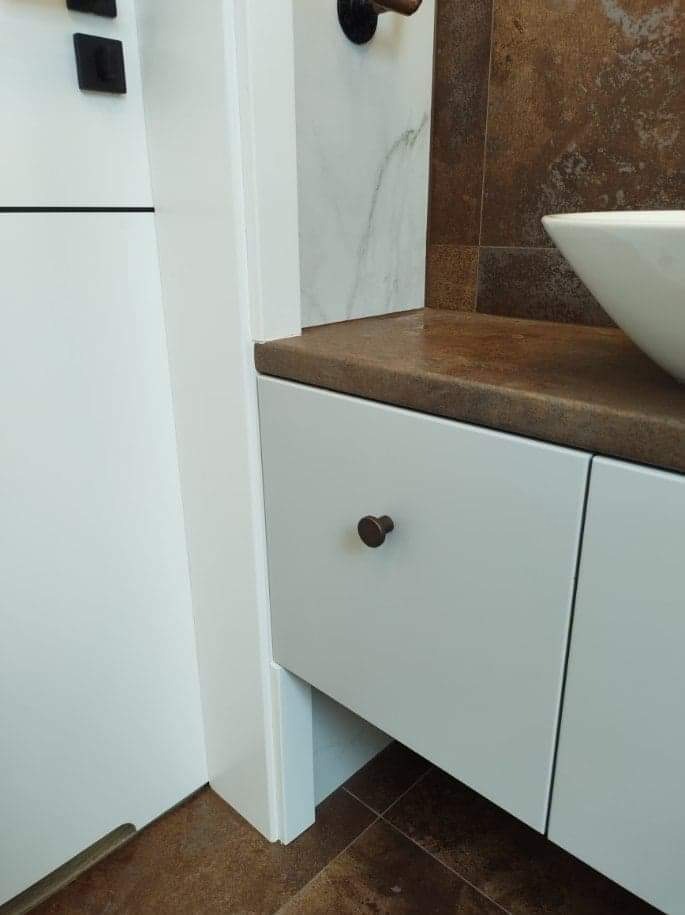 Fragment łazienki z umywalką nablatową, białą szafką z brązowymi uchwytami i brązową płytką imitującą rdzę na ścianie oraz podłodze.