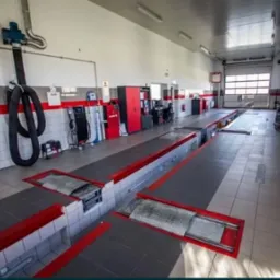 Wnętrze warsztatu samochodowego z kanałami naprawczymi, widoczne urządzenia diagnostyczne i wentylacja przemysłowa.