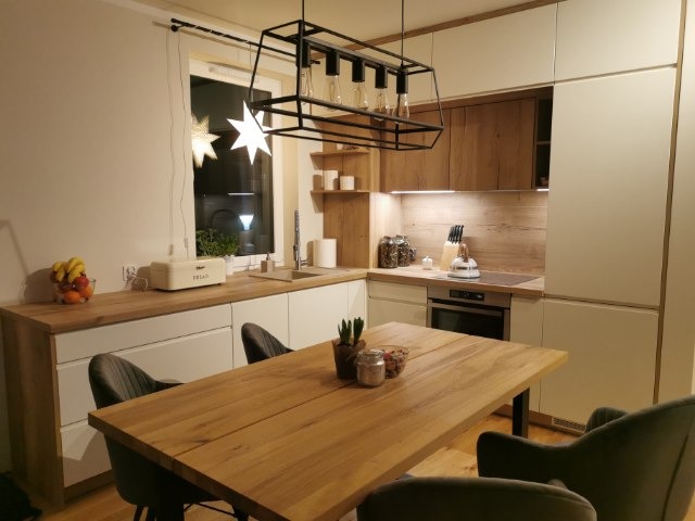 Nowoczesna kuchnia z drewnianymi blatami i frontami szafek, oświetlona lampą w stylu industrialnym, stół z krzesłami w odcieniach szarości, dekoracje świąteczne w oknie.