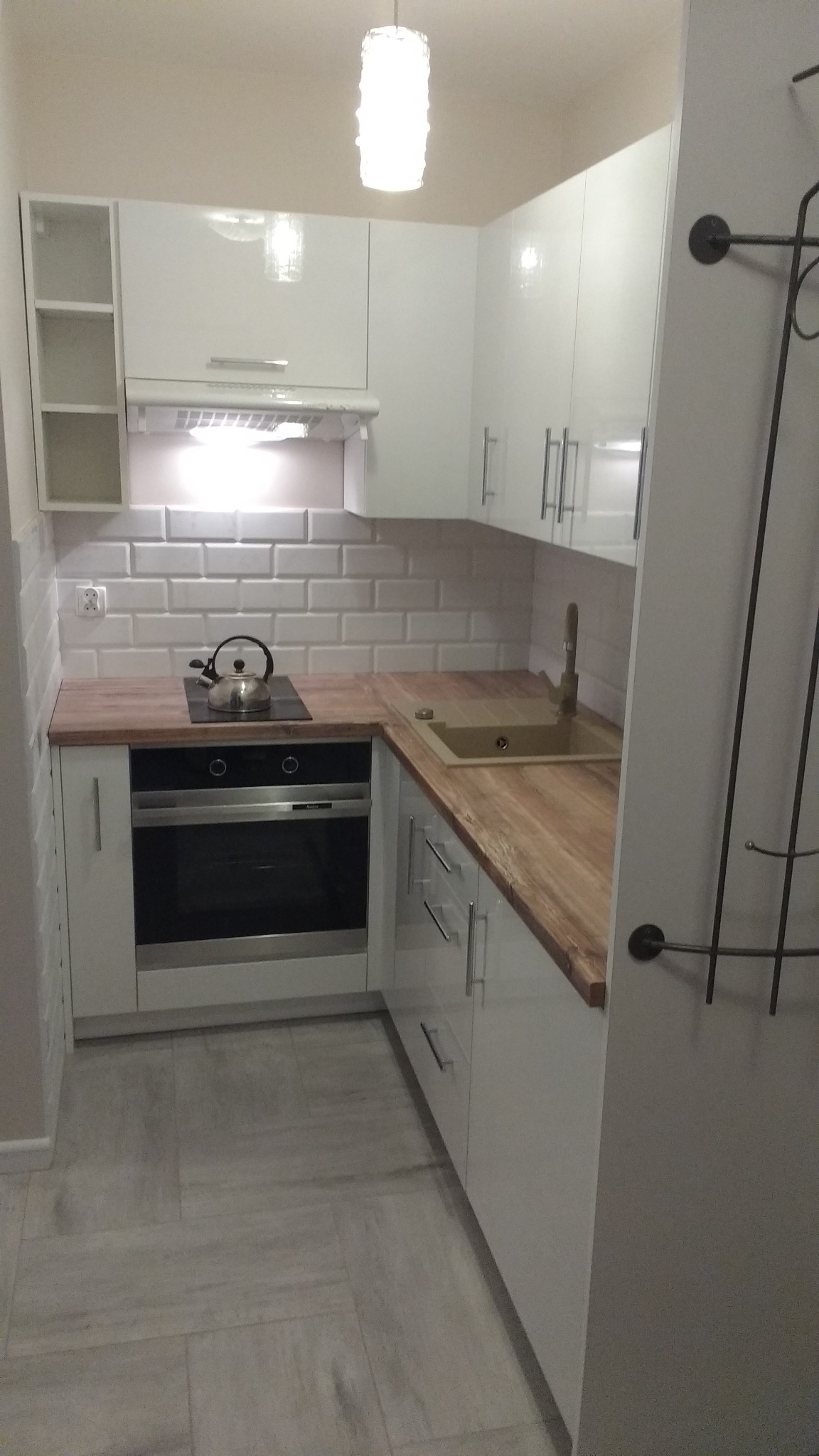 Nowoczesna, jasna kuchnia z białymi, lakierowanymi szafkami, drewnianym blatem i płytkami typu metro. Na blacie czajnik na płycie indukcyjnej, oświetlenie podszafkowe.
